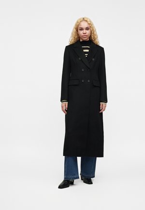 Mujer con cabello largo y rubio que lleva un abrigo largo negro de doble botonadura, suéter de rayas, vaqueros anchos azules y zapatos negros, de pie sobre un fondo blanco.