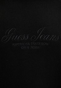 Tessuto nero con una texture liscia presenta un logo ricamato con la scritta "Guess Jeans" sopra "American Tradition California" in un design sottile.