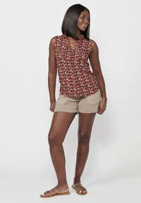 Blusa sin mangas roja con flores y cuello, combinada con pantalones cortos beige que tienen bolsillos laterales y una textura suave, usada con sandalias planas.