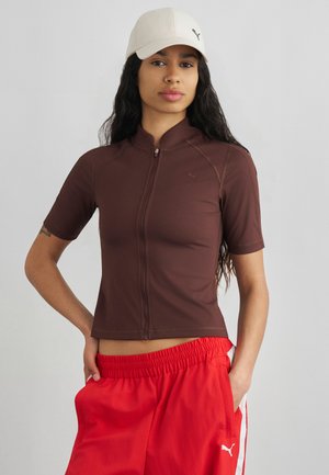 Ung kvinde med langt mørkt hår iført en hvid kasket, brun kortærmet lynlås-top og røde elastiske bukser med hvid stribe i taljen.