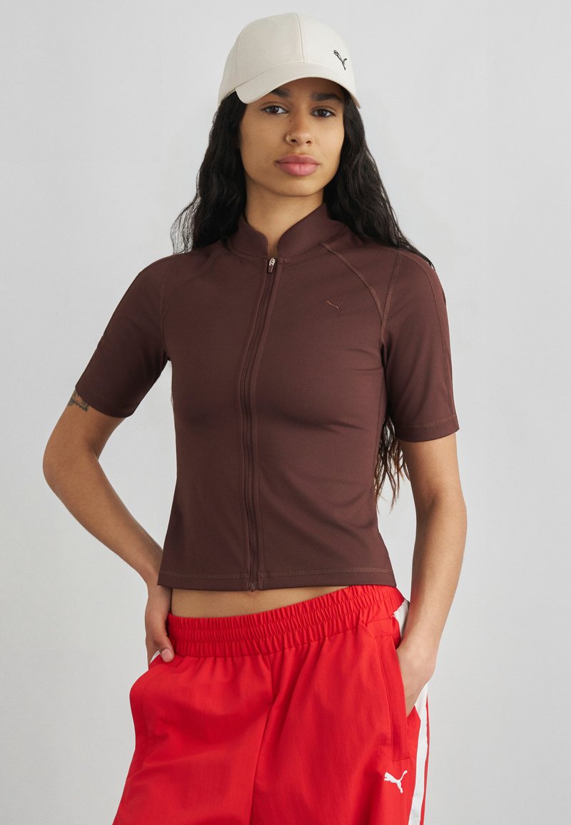 Νεαρή γυναίκα με μακριά σκούρα μαλλιά που φοράει λευκό καπέλο, καφέ τοπ με κοντά μανίκια και φερμουάρ, και κόκκινο παντελόνι με ελαστική μέση και λευκή ρίγα.