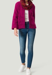 Fuchsiová texturovaná bunda s širokými rukávy a dvěma předními kapsami, spárovaná s modrými skinny džíny a bílými teniskami, doplněná vzorovaným šátkem.