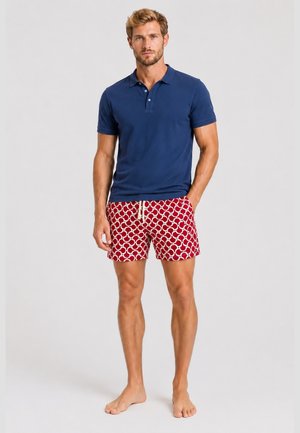Ein Mann trägt ein marineblaues Poloshirt und rote gemusterte Badeshorts und steht barfuß vor einem einfarbigen hellen Hintergrund.