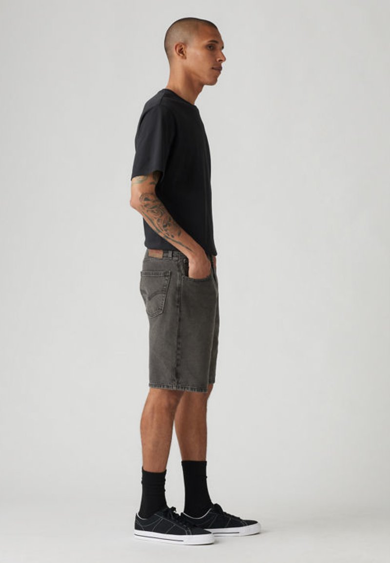 Mand med tatoveret arm iført sort t-shirt, grå denimshorts, sorte sokker og sorte sneakers, stående i profil med hænderne i lommerne.