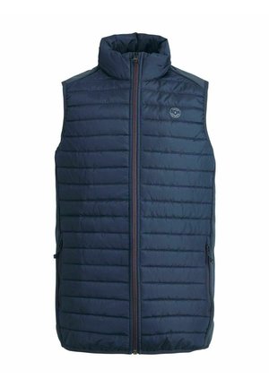 Person iført en marineblå quiltet vest over en grå sweater med sorte og hvide striber, blå jeans og sorte sneakers med hvide snørebånd.