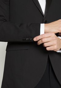 Giacca nera con una texture liscia, bavero strutturato e quattro bottoni neri sul polsino. Polsino della camicia bianca visibile sotto.