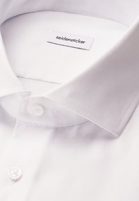 Collo e primi due bottoni di una camicia bianca, che mostrano la texture del tessuto e un'etichetta "seidensticker" all'interno del collo.