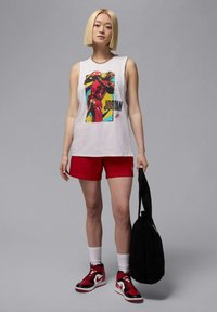 Camiseta sin mangas blanca con un gráfico colorido de Michael Jordan, combinada con pantalones cortos rojos y zapatillas negras. Sosteniendo una bolsa de tote negra.