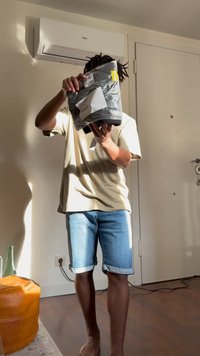 Άτομο που φοράει μπεζ μπλουζάκι και τζιν σορτς κρατά μια σφραγισμένη μαύρη πλαστική συσκευασία σε εσωτερικό χώρο κοντά σε λευκή πόρτα και κλιματιστικό.