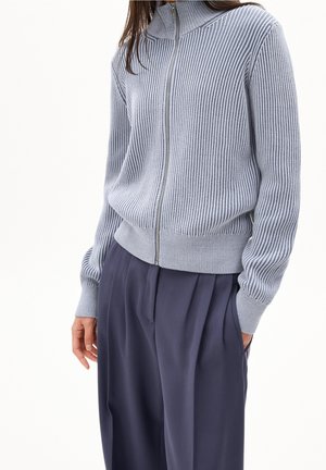 Kvinde iført lyseblå ribstrikket lynlås-sweater og mørkeblå plisserede bukser med den ene hånd i lommen mod hvid baggrund.