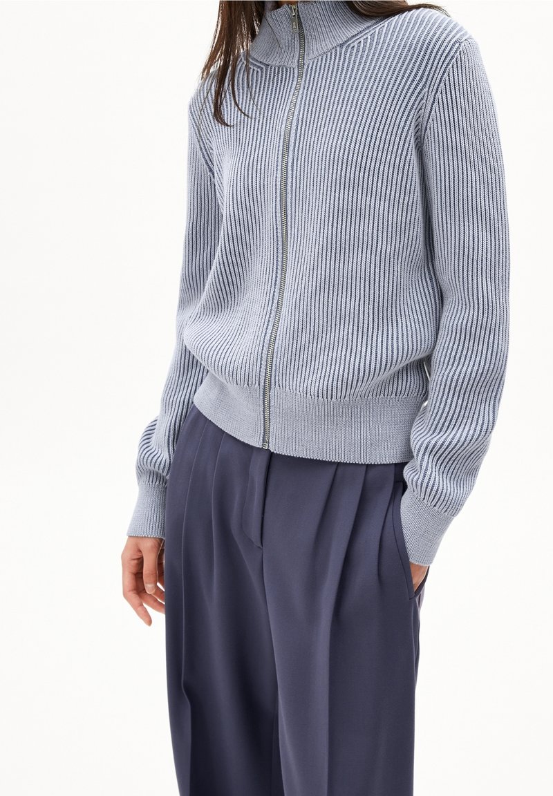 Femme portant un pull zippé côtelé bleu clair et un pantalon plissé bleu foncé, avec une main dans la poche, sur fond blanc.