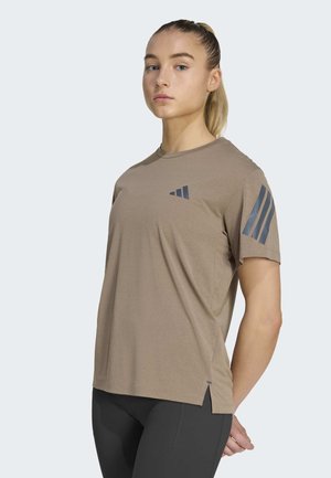 Junge Person mit zurückgebundenem Haar, trägt ein braunes Sport-T-Shirt mit dunklen Streifen auf Ärmeln und Brust, kombiniert mit schwarzen Leggings.