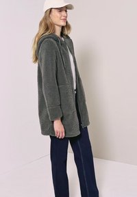 Vrouw die een grijs fleecejack met capuchon, witte pet, wit shirt en donkere wijde broek draagt, staand tegen een effen lichte achtergrond.