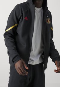 Svart zip-up jacka med gul rand och röd Adidas-logga; har Atlanta United SC-emblemet på bröstet; tillverkad av ett slätt material.