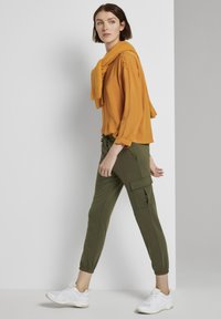 Camicia a maniche lunghe gialla senape con polsini arrotolati, abbinata a pantaloni cargo verde oliva con tasche laterali, completata da sneakers bianche.