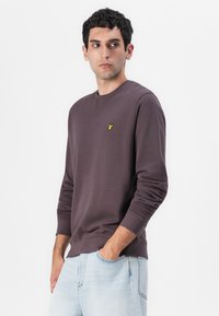 Mørkebrun sweatshirt laget av bomullsblanding med lange ermer, rund hals, og en liten gul logo på venstre bryst. Lys blå jeans.