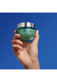 Grønn glasskrukke med sølvlock, merket "Biotherm Aquasource Hyalu Plump Gel," holdt i en hånd mot en blå bakgrunn.