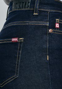 Sötétkék farmernadrág közeli kép sárga varrással, piros szövetcímkékkel, amelyeken fehér csillag látható, és fekete bőr márkajelzéssel a derékrészen.