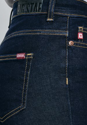 Blugi din denim albastru închis, detaliu apropiat, cu cusături galbene, etichete din material roșu cu o stea albă și o etichetă din piele neagră cu brand pe talie.