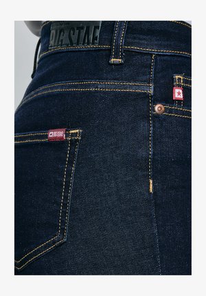 Primer plano de jeans de mezclilla azul oscuro con costuras amarillas, etiquetas de tela rojas con una estrella blanca y una etiqueta de cuero negro con la marca en la cintura.