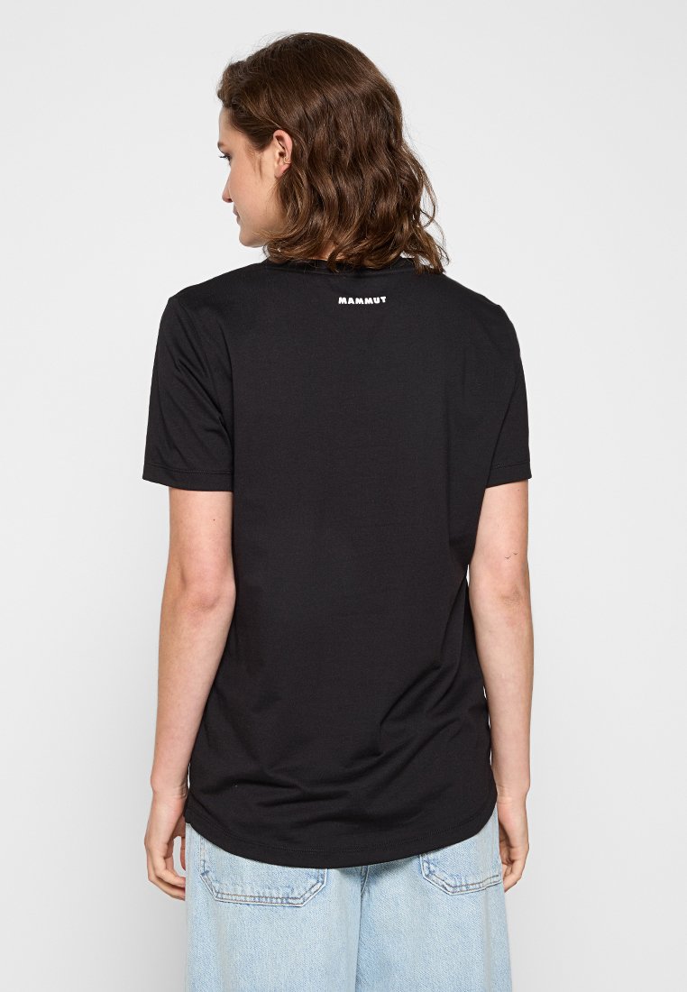 Person mit schulterlangem braunem Haar trägt ein schwarzes Mammut-T-Shirt und hellblaue Jeans, von einer schlichten weißen Hintergrund abgewandt.