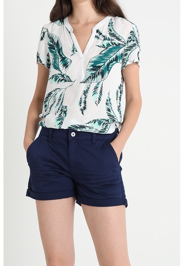 Femme portant un chemisier blanc à manches courtes avec un imprimé de feuilles vertes et un short bleu marine à ourlet roulé, les mains dans les poches.