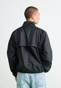 Mand med kort hår iført en sort vindjakke og lyseblå denim jeans, der står med ryggen til en ensfarvet hvid baggrund.