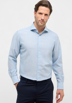 Uomo che indossa una camicia azzurra a maniche lunghe infilata in pantaloni scuri, in piedi con una mano in tasca.