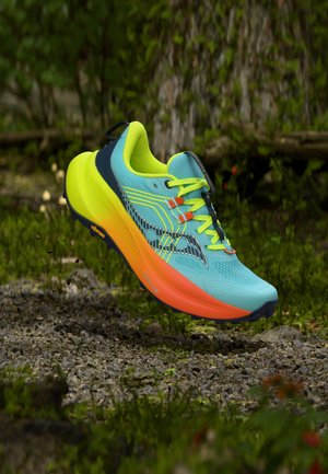 Heldere turquoise en oranje hardloopschoen met neon gele veters zwevend boven een grind- en mosachtig bosgrond.