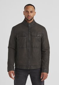 Dunkle Lederjacke mit hohem Kragen, Frontreißverschluss und zwei Brusttaschen. Glatte Oberfläche und taillierte Passform, kombiniert mit dunklen Jeans.