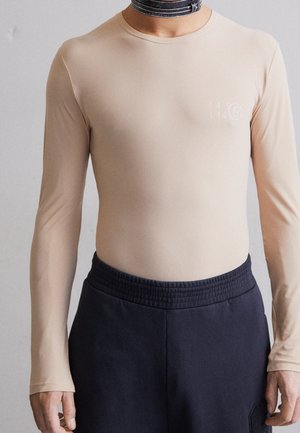 Personne portant une chemise beige ajustée à manches longues avec un imprimé discret sur la poitrine et un pantalon noir à taille élastique sur un fond uni.