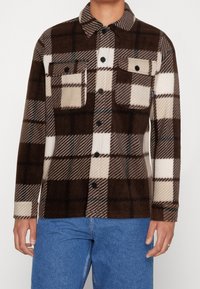 Chaqueta de cuadros en marrón y crema hecha de forro polar, con dos bolsillos en el pecho, botones negros y un diseño de cuello clásico.