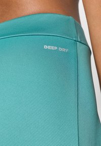 Tirkizna sportska majica s teksturiranom površinom. Na oznaci piše "DEEP DRY" u bijelom, što označava materijal koji odvlači vlagu. Mogu se vidjeti detalji šavova.