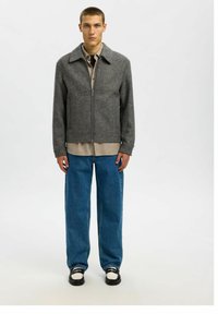 Siva herringbone jakna na zip z izrezanim ovratnikom, nošena čez bež srajco, v kombinaciji z ohlapnimi modrimi jeans hlačami in črnimi ter belimi čevlji.