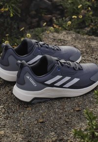 Paire de chaussures de trail Adidas Terrex grises et noires avec des accents rayés blancs reposant sur du gravier en plein air près de petites fleurs jaunes.