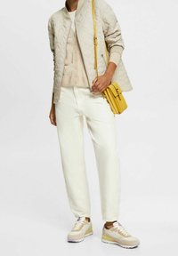 Osoba w beżowej pikowanej kurtce, jasnobeżowym swetrze, off-white spodniach, beżowych trampkach z fioletowymi akcentami, trzymająca żółtą torbę na ramię.