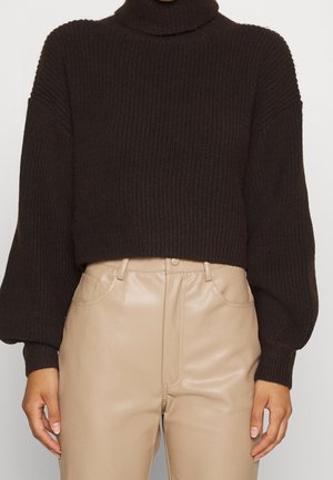 Gros plan sur une personne portant un pull à col roulé côtelé marron foncé et un pantalon en cuir beige taille haute avec des poches avant.