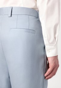 Pantalones de color azul claro hechos de una tela texturizada, con un bolsillo trasero plano y un corte entallado, combinados con una camisa blanca de botones.