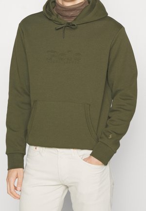 Man draagt een kaki groene hoodie met kangoeroezak en geperst "Polo Ralph Lauren" logo, gecombineerd met licht beige broek.