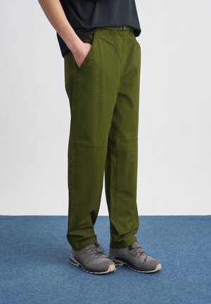 Άτομο που φοράει ελιά πράσινο παντελόνι με ελαστική μέση και γκρι παπούτσια πεζοπορίας, στέκεται σε μπλε χαλί.