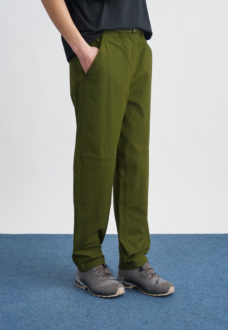 Άτομο που φοράει ελιά πράσινο παντελόνι με ελαστική μέση και γκρι παπούτσια πεζοπορίας, στέκεται σε μπλε χαλί.