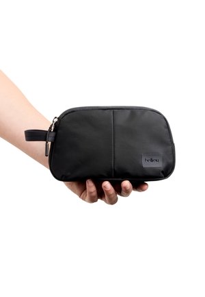 Mano che tiene una piccola pochette nera con cerniera, dotata di cinturino da polso e logo "bellroy" sul davanti.