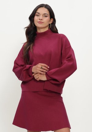 Woman wearing a matching dark pink sweater with balloon sleeves and a skirt, standing with hands clasped and neutral expression.