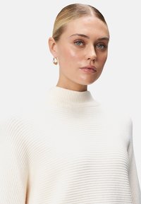 Pull en maille côtelée de couleur crème avec un col montant et des manches longues, présentant une texture douce. De simples boucles d'oreilles en or ajoutent une touche à l'ensemble.