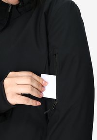 Giacca impermeabile nera con tasca laterale con zip; una mano sta inserendo una scheda bianca nella tasca. Texture liscia, design minimalista.