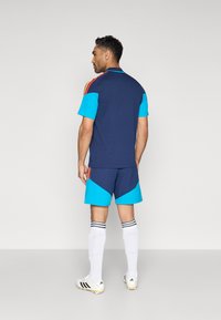 Ανδρική αθλητική εμφάνιση με μπλε σκούρο τοπ με μπλε μανίκια και κόκκινες ρίγες, σε συνδυασμό με μπλε σορτς, λευκές κάλτσες και παπούτσια ποδοσφαίρου.