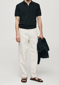 Polo noir à manches courtes, pantalon beige à plis, sandales marron. Tenant une veste sombre. Tissu lisse, coupe décontractée, design minimaliste.