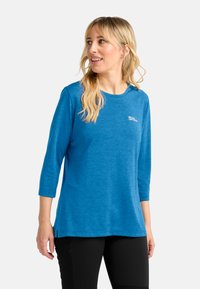 Vrouw met blond haar, gekleed in een blauwe longsleeve shirt en zwarte broek, kijkt naar links tegen een plain witte achtergrond.