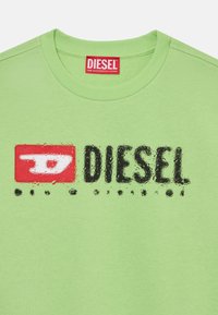 T-shirt in cotone verde con colletto rotondo, logo grafico testurizzato in rosso e nero, e un'etichetta con "DIESEL" in lettere maiuscole.