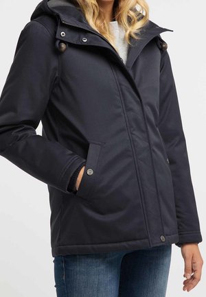 Nainen pukeutuneena tummansiniseen hupulliseen talvitakkiin, jossa on vetoketju ja nepparitaskut, yhdistettynä sinisiin farkkuihin, seisomassa yhdellä kädellä taskussa.